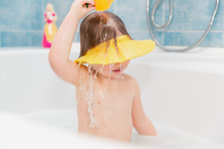 A toddler wearing a yellow bath visor and pouring water over her head.