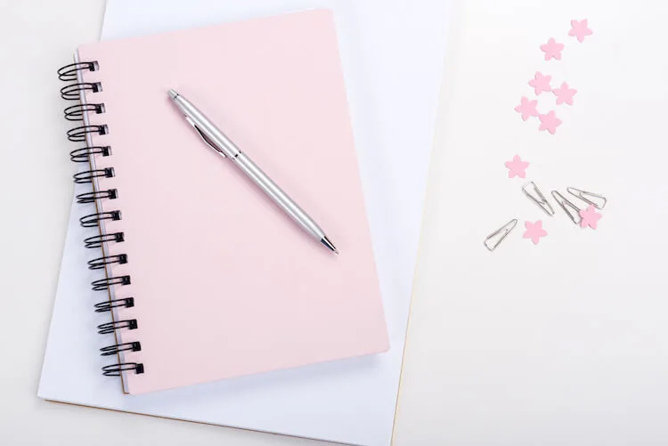 A pink journal lays on top of a white notepad. There's a pen on top of it and some pink confetti on the white desk beside it.
