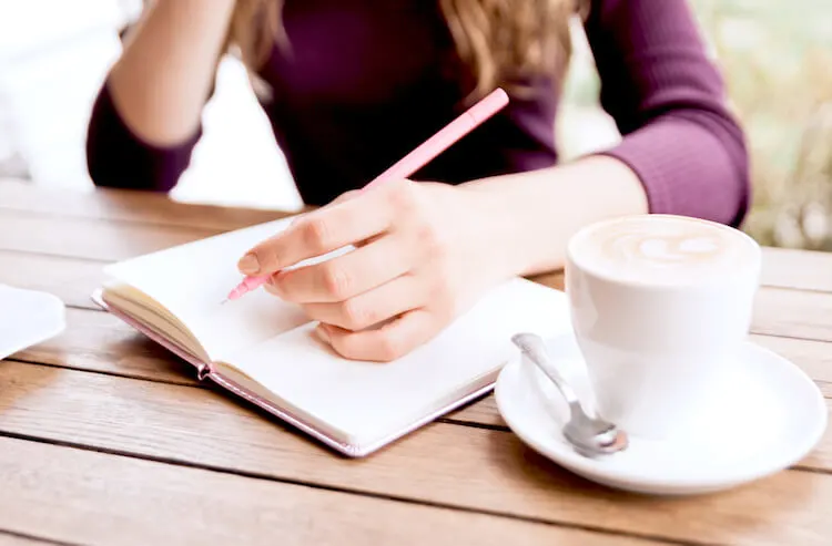 A woman is writing in a journal on a slatted wooden table top. She wears a dark purple jumper and holds a poised pen. She has a white mug beside her.