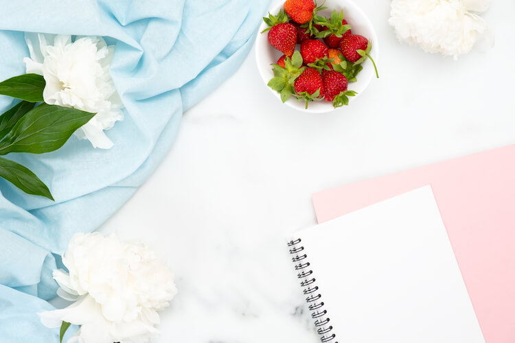 Open notebook with blue scarf and white flowers and a bowl of strawberries in background.