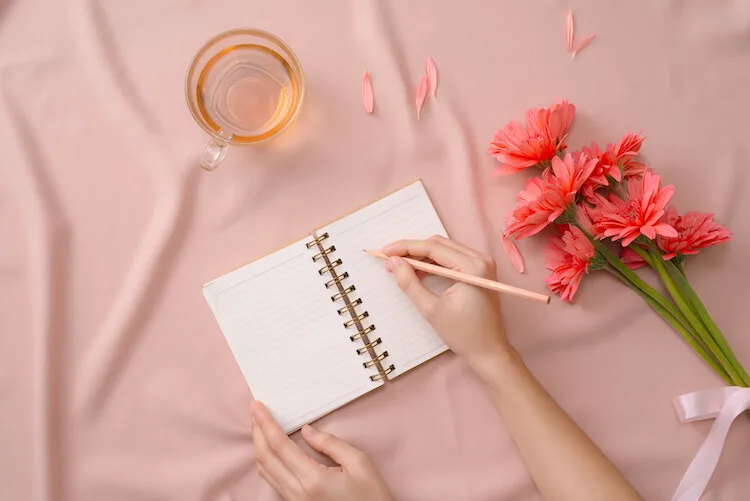 How to start journaling | Journaling tips | Image shows a woman writing in a journal with a peach background and a cup of tea beside her.