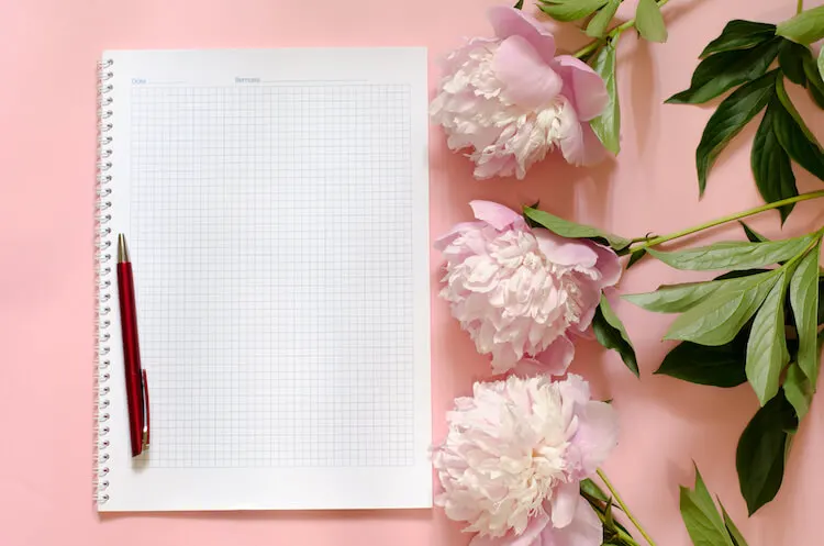 How to write a journal | Image shows a journal and pen on a pink background beside some pink peonies.