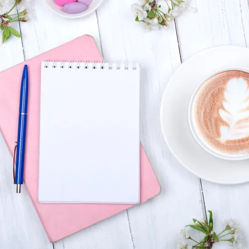 Journaling ideas | Image shows a journal and pen on a white desk beside a cup of coffee and some flowers.