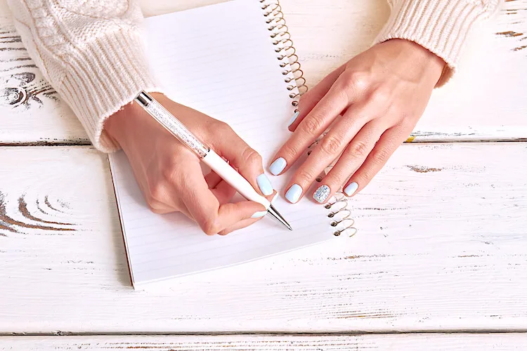 Image shows a woman writing in a notebook. She wears pale blue nail varnish and a creamy jumper.