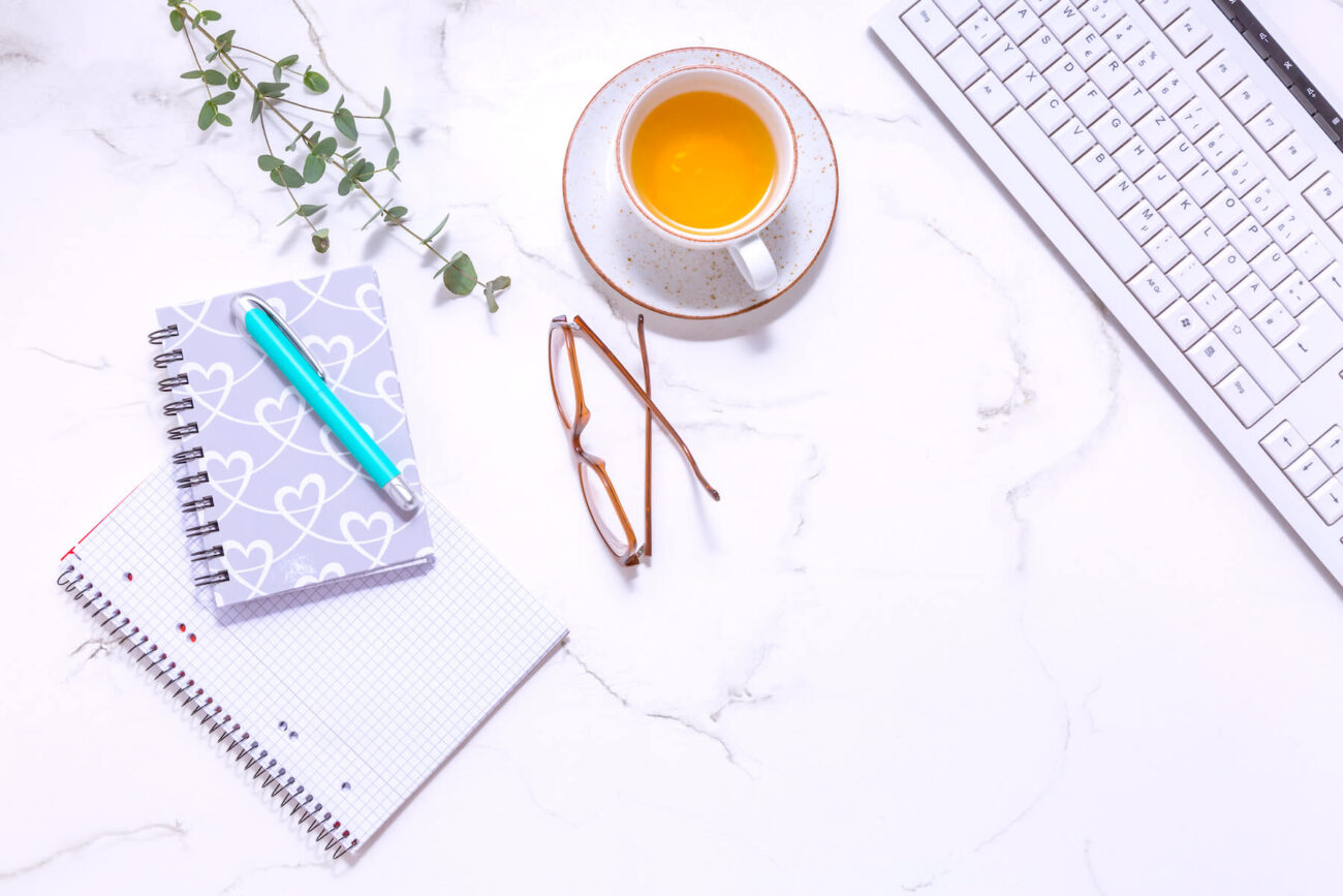 Journal prompts for beginners | A white desk holding a notebook and pen on top of a journal, a pair of reading glasses, a keyboard and a cup of herbal tea.