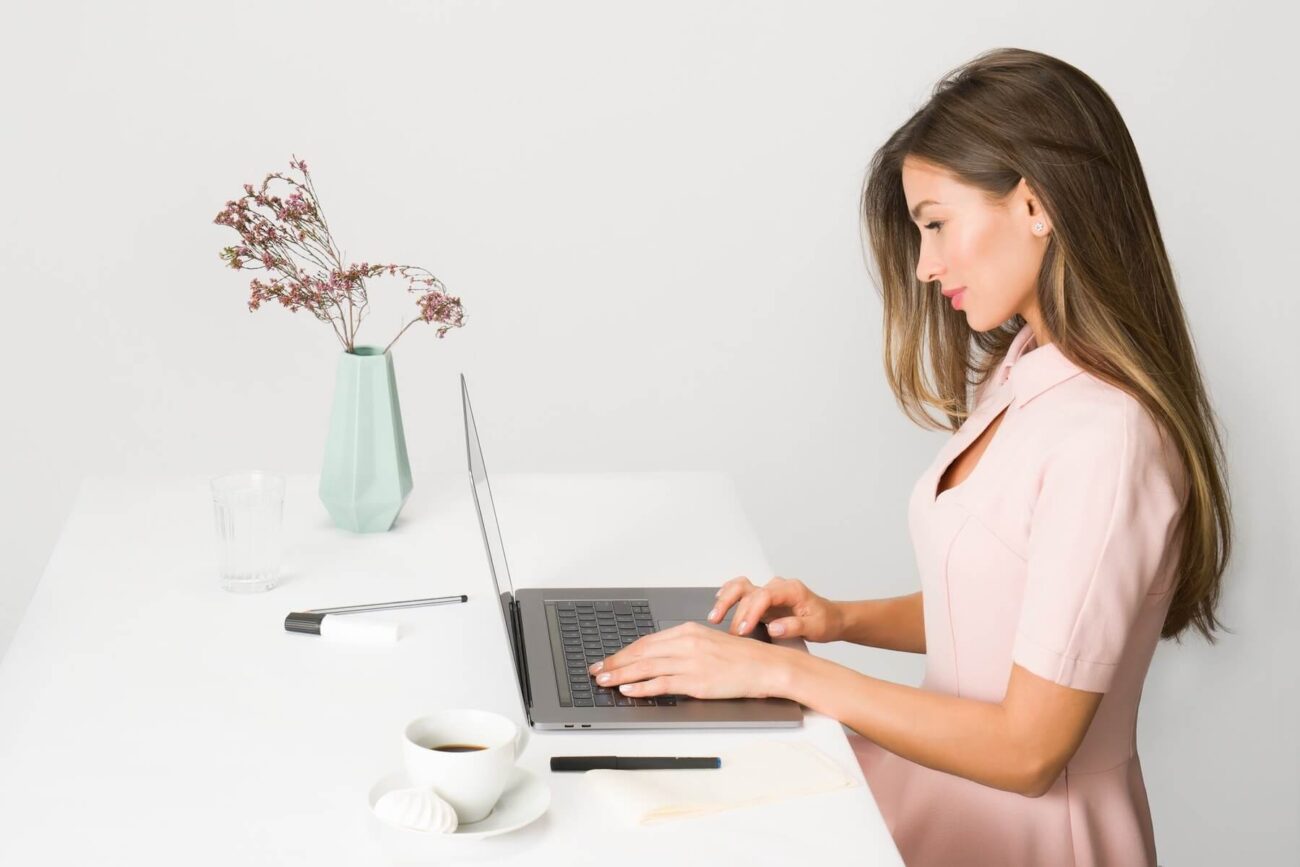 Female business headshot of a woman typing at a desk. There's a green vase of flowers beside her.