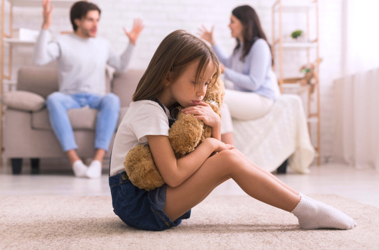 Co-parenting with a narcissist | A young girl sits in the foreground cuddling her teddy, with her parents arguing on the sofa behind her.