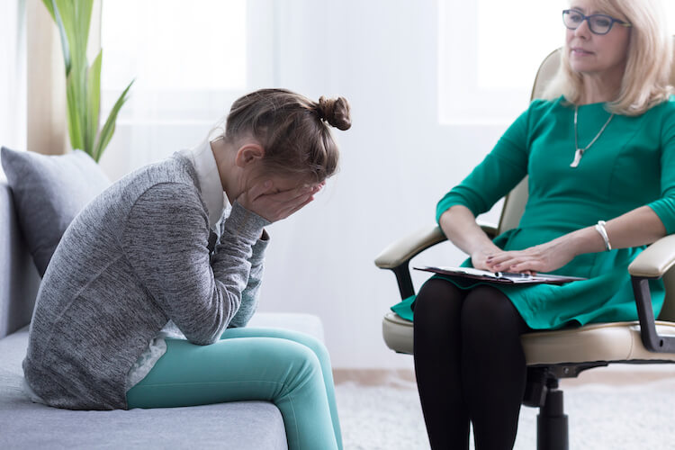 A girl wearing a grey cardigan and aqua coloured trousers sits in front of her therapist with her head in her hands.