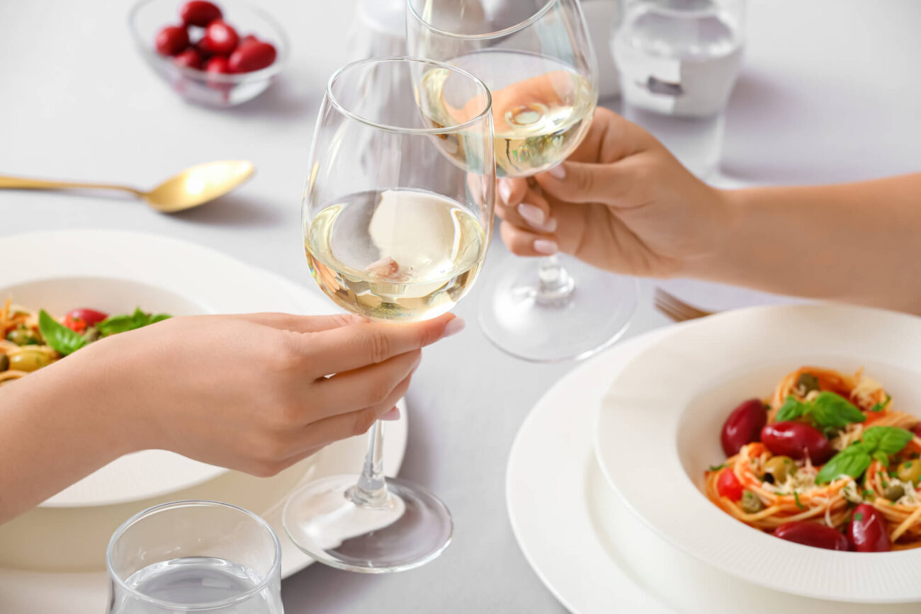 Two women clinking wine glasses across a restaurant table. There are plates of colourful food in the background.