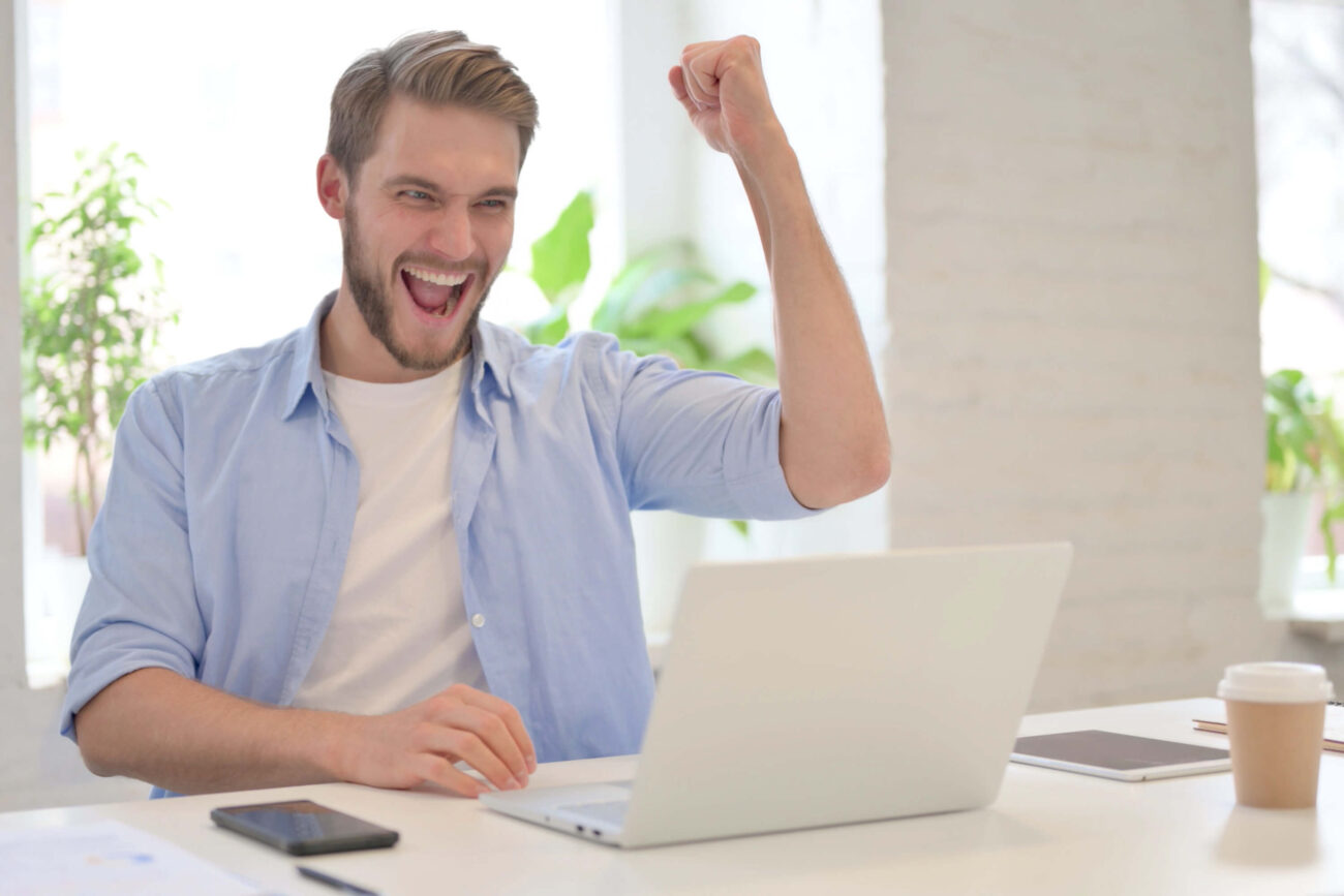 Goal setting | A man in a blue sheet celebrates success with a fist pump. He sits at a desk in front of his laptop.