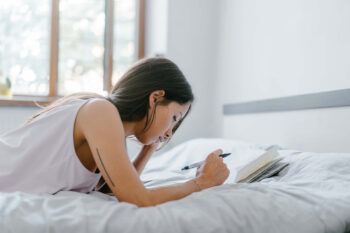 Journal prompts for teens | A teenage girl with dark hair lays on her bed writing in her journal.