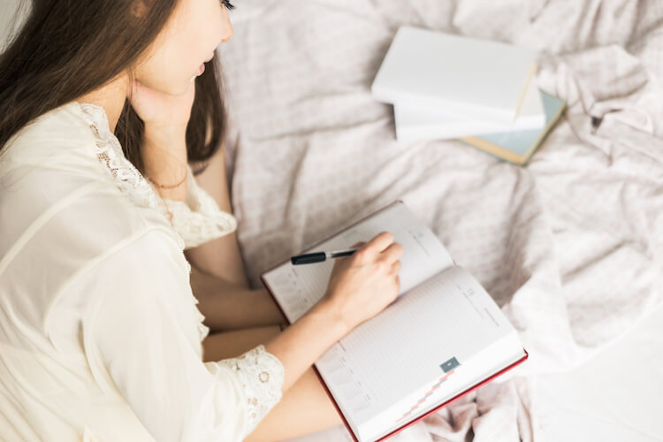 A teenage girl with long dark hair and wearing a white cardigan sits on her bed with her journal on her knees and a pen in her hand.