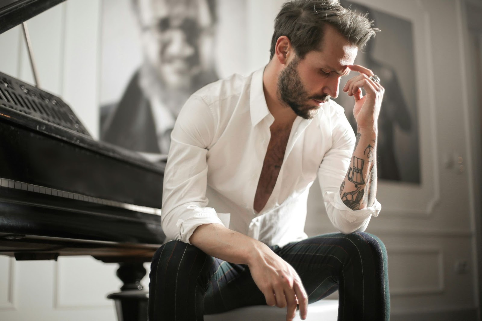 A man sits beside a piano with his hand to his head. He's wearing a white shirt and dark trousers.